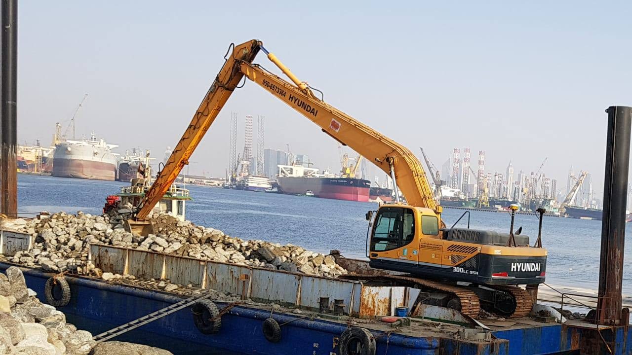 MACO Hyundai long-boom excavator on a barge with cargo ships and rigs at Jebel Ali Port