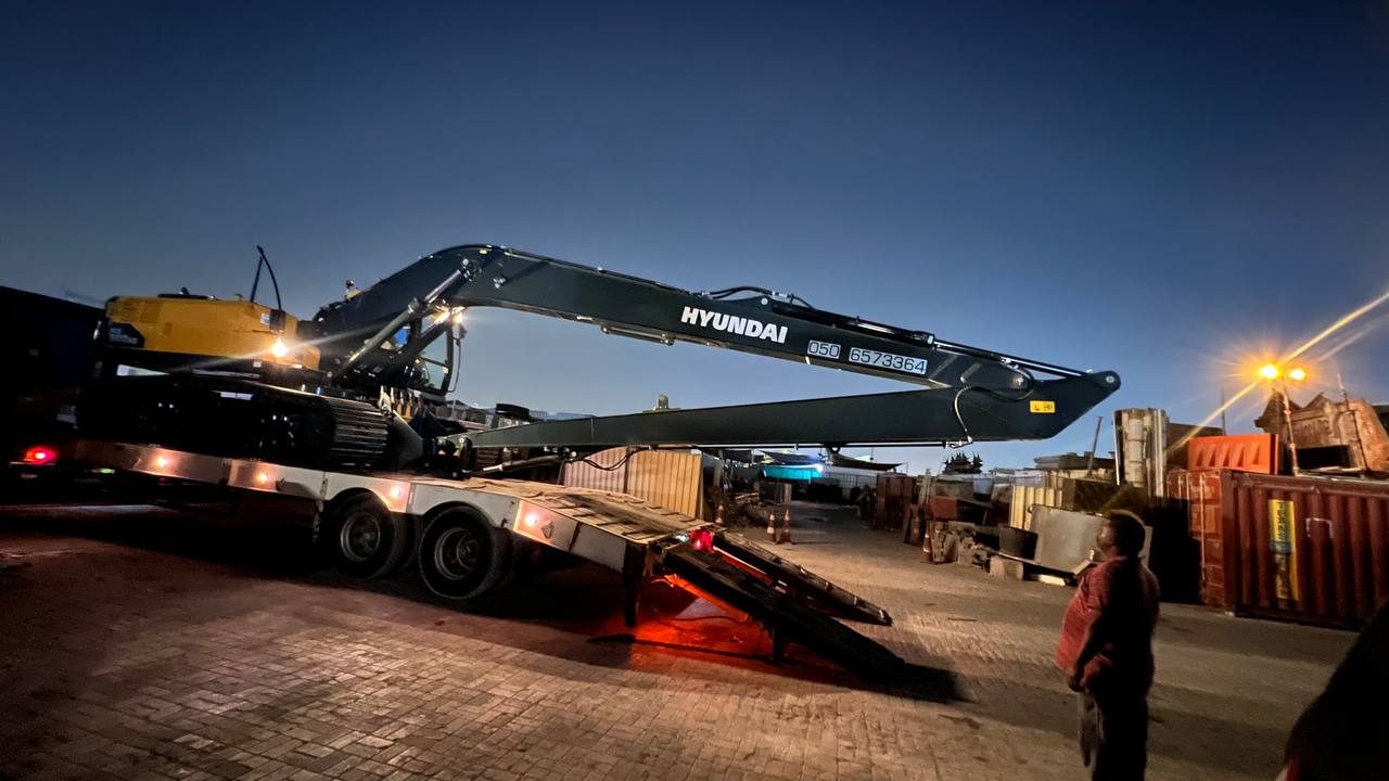 MACO Hyundai long-boom excavator loaded on a low-bed trailer at dusk